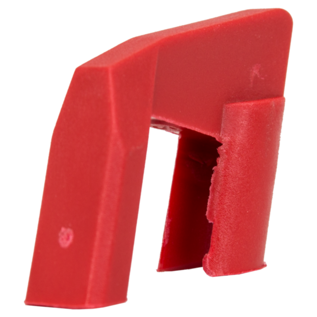 A red, angular plastic item similar to the MAGAZINE FOLLOWER KADET, featuring a narrow opening in the center and an uneven surface. It has a slanted top and asymmetrical sides, akin to other Kadett accessories.