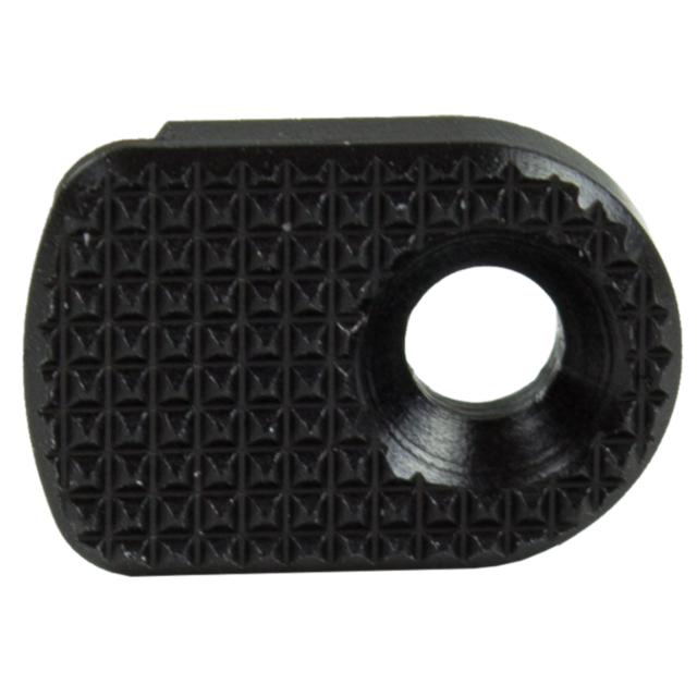 A close-up of the MAGAZINE CATCH PADDLE SHADOW 2 BLACK, a small, black, textured metal plate, designed with an oval shape and a circular hole on one side for potential mechanical use. Its surface is adorned with a grid of raised, diamond-shaped patterns.