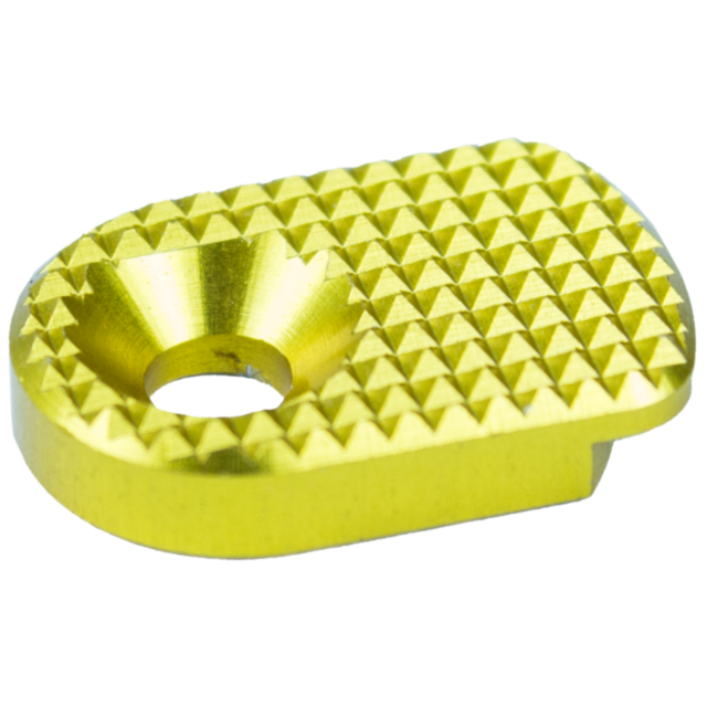 The image shows a detailed view of the MAGAZINE CATCH PADDLE SHADOW 2 GOLD, a small, glossy gold piece with a circular hole in the center. Its top surface is adorned with a textured diamond pattern, and it has rounded edges.