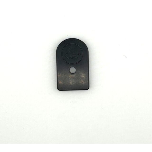 The DWX COMPACT MAGAZINE BASE PAD BLACK is a small, rectangular, dark metal object with a rounded top, engraved circular design at the top, and a central hole. Its surface is smooth and reflective.